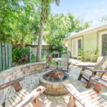 Backyard area with a fire pit surrounded by Adirondack chairs, lush greenery, and a patio with outdoor seating at a vacation rental.