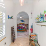 A hallway with bright natural light coming through plantation shutters, featuring coastal-themed artwork and decor. An arched doorway reveals a vibrant, tropical-themed bedroom with a hand-painted bed and colorful oceanic decor, embodying a warm and welcoming coastal atmosphere.