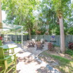 Backyard patio area with a fire pit, Adirondack chairs, and a green bistro table set, surrounded by greenery and tall trees, offering a peaceful retreat at a vacation rental.