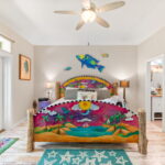 A vibrant bedroom with a hand-painted bed featuring tropical scenes, colorful fish wall art, and coastal-themed decor. The room is bright with natural light from a large window and glass door, creating a serene and whimsical atmosphere.