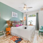 A cozy bedroom with a coastal theme, featuring a colorful headboard, vibrant pillows, and ocean-inspired decorations. The room has a ceiling fan, wooden flooring, and a large window letting in natural light. A small bench and wooden ladder add to the beachy vibe.