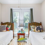 A twin bedroom with two beds dressed in white linens and colorful, tropical-themed pillows. The room features a turtle-themed rug and wall decor, striped headboards, and French doors that open to a lush garden, allowing natural light to fill the space. The room has a bright and cheerful atmosphere, perfect for relaxation.