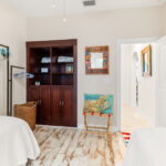 A twin bedroom with two beds dressed in white linens, featuring a built-in wooden cabinet, a coastal-themed cushion on a small chair, and a mirror on the wall. The room has neutral tones with coastal decor accents, creating a cozy and welcoming atmosphere. A doorway leads to the main living areas of the home.