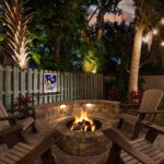 Outdoor fire pit area at St. Augustine Guest House with Adirondack chairs, surrounded by trees and illuminated by string lights and landscape lighting.