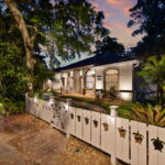 Front view of a house at dusk with warm exterior lighting, surrounded by trees and a white fence with sea turtle cutouts, creating a welcoming and tranquil atmosphere.