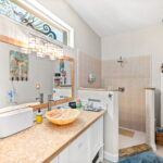 A well-lit bathroom with a coastal theme, featuring a glass vessel sink on a mosaic countertop, a large mirror with natural wood framing, and a walk-in shower. The room is adorned with nautical decorations, including a vibrant mermaid wall art.