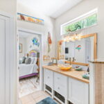 A well-lit bathroom with a coastal theme, featuring a glass vessel sink on a mosaic countertop, a large mirror with natural wood framing, and a walk-in shower. The room is adorned with nautical decorations, including a vibrant mermaid wall art.